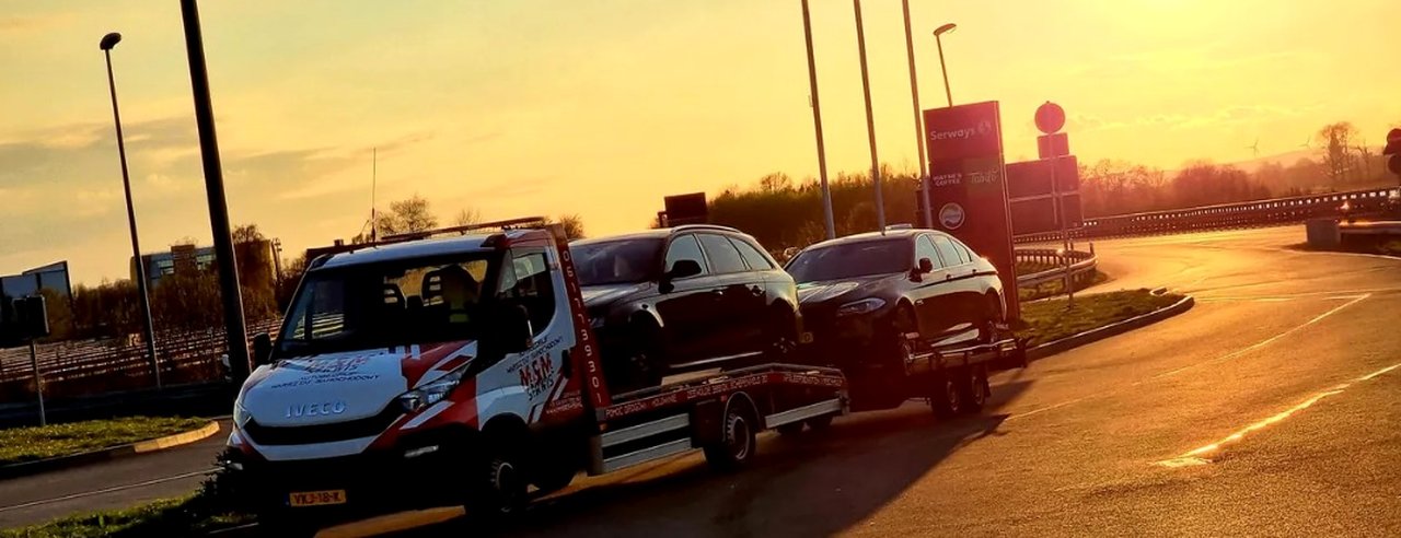 Iveco bergingswagen met twee auto's, één op de laadbak en één op de aanhanger, bij zonsondergang